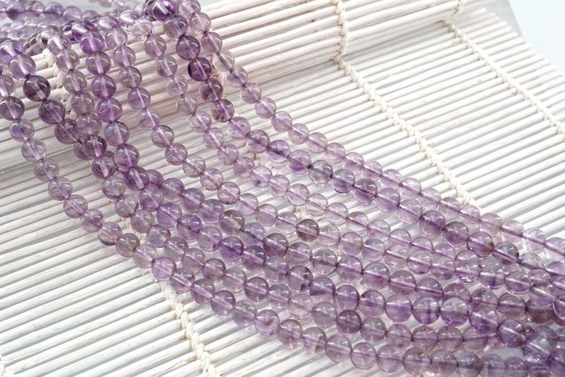 May include: A strand of amethyst beads, with a light purple color, are arranged in rows on a white woven surface. The beads are round and smooth.