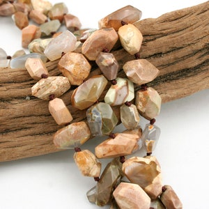 May include: A strand of brown, beige, and white gemstone beads with a faceted, irregular shape. The beads are strung on a brown cord and are displayed on a piece of driftwood.