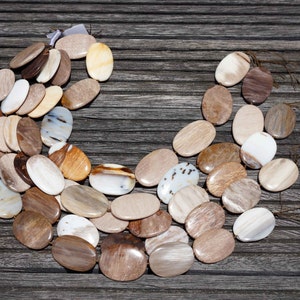Puede incluir: Un collar hecho de cuentas ovaladas de madera petrificada en tonos marrones, beige y blancos. Las cuentas est&aacute;n dispuestas en un patr&oacute;n curvo, creando una forma de media luna.