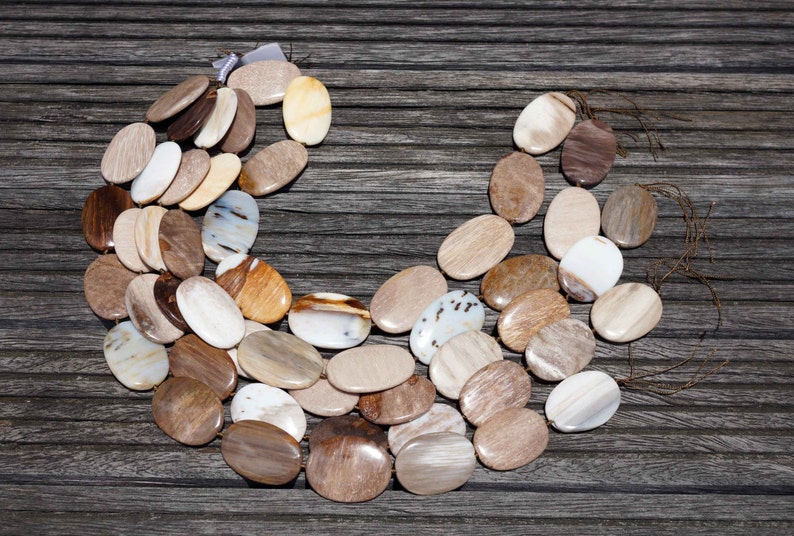 Puede incluir: Un collar hecho de cuentas ovaladas de madera petrificada en tonos marrones, beige y blancos. Las cuentas est&aacute;n dispuestas en un patr&oacute;n curvo, creando una forma de media luna.