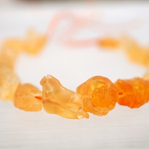 May include: A close-up of a necklace made of orange amber beads. The beads are irregular in shape and have a translucent quality. The necklace is lying on a white surface.