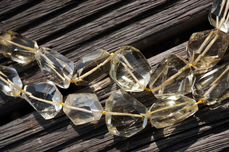 May include: A strand of clear, faceted gemstone beads with a golden yellow hue. The beads are strung on a thin, gold wire and are arranged in a staggered pattern. The beads are lying on a dark wooden surface.