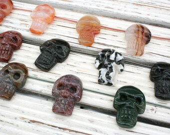 May include: A collection of 11 hand-carved skull shaped crystal pendants in various colors and patterns, including black, white, red, green, and brown. The pendants are arranged on a white wooden surface.