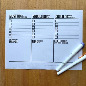 May include: A white paper checklist with the headings "Must Do," "Should Do," and "Could Do." The checklist includes sections for errands, fun, and things to buy. Two white pens with colored caps rest on the paper.