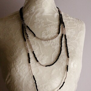May include: A layered necklace with black and light pink beads. The necklace is displayed on a white lace mannequin. The beads are small and evenly spaced, creating a delicate and elegant look. The necklace is a versatile accessory.