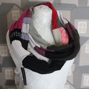 Może przedstawiać: Wielokolorowy szal typu infinity wykonany z recyklingowanych swetrów. Szal jest wykonany w różnych odcieniach różowego, szarego, czarnego i czerwonego. Szal jest prezentowany na białym manekinie z koronkowym wzorem. Szal ma metkę z napisem "Anna's 7 Creations".
