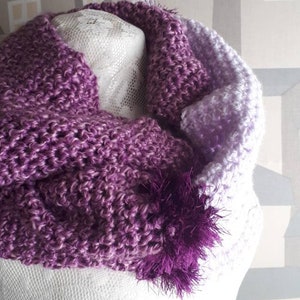 May include: A purple and lavender knitted scarf with a fluffy purple pom-pom. The scarf is made of a soft, chunky yarn and has a gradient color pattern.