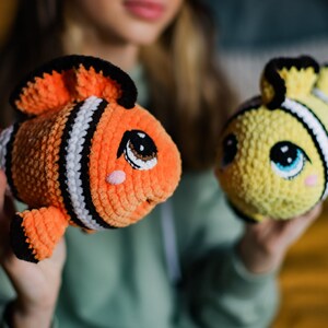 May include: Two crocheted stuffed fish toys. One is orange and black with white stripes and the other is yellow with black stripes. Both have big eyes and a cute expression.