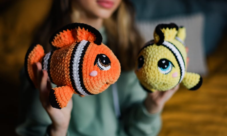 May include: Two crocheted stuffed fish toys. One is orange and black with white stripes and the other is yellow with black stripes. Both have big eyes and a cute expression.