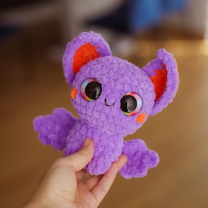 Puede incluir: Un murci&eacute;lago de crochet morado con orejas naranjas y ojos grandes y coloridos. El murci&eacute;lago est&aacute; siendo sostenido por una mano.