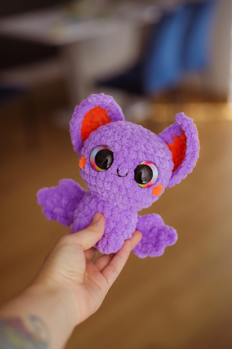 Puede incluir: Un murci&eacute;lago de crochet morado con orejas naranjas y ojos grandes y coloridos. El murci&eacute;lago est&aacute; siendo sostenido por una mano.