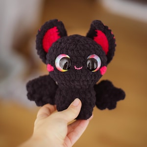 Puede incluir: Un peque&ntilde;o murci&eacute;lago de crochet negro con orejas rojas y grandes ojos arco&iacute;ris. El murci&eacute;lago sonr&iacute;e y tiene una nariz rosa.