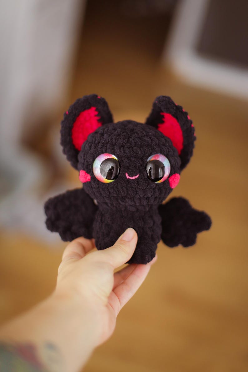Puede incluir: Un peque&ntilde;o murci&eacute;lago de crochet negro con orejas rojas y grandes ojos arco&iacute;ris. El murci&eacute;lago sonr&iacute;e y tiene una nariz rosa.