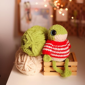 Peut inclure: Une grenouille au crochet verte et blanche faite à la main porte un pull rayé rouge et blanc et est assise sur une petite caisse en bois. Deux pelotes de laine, une vert clair et une beige, sont à côté de la grenouille. La grenouille a de grands yeux bleus.