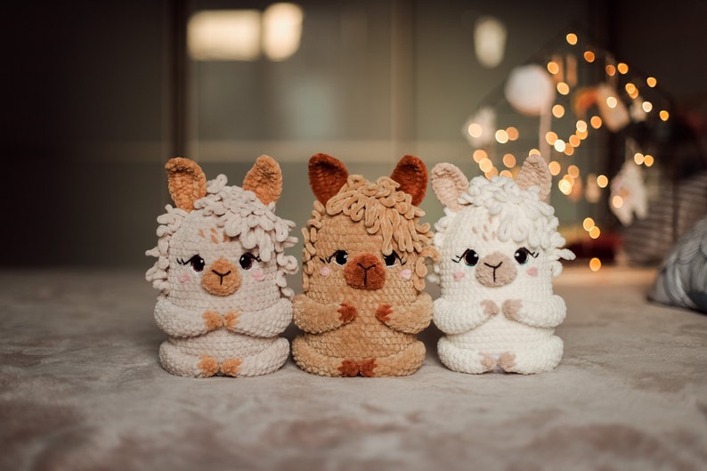 May include: Three handmade crochet llama dolls in cream, tan, and brown. Each llama has a fluffy head, small ears, and a sweet face with closed eyes. The dolls are sitting side-by-side.