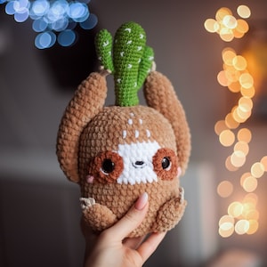 Puede incluir: Un juguete de peluche de perezoso tejido a crochet con un cactus verde en la cabeza. El perezoso es marrón con una cara blanca y ojos negros.