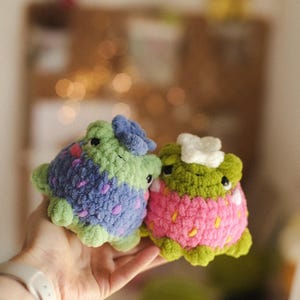 May include: Two handmade crochet frog plushies. One frog is green and blue with purple accents, the other is green and pink, resembling a strawberry. The plushies are held in a hand, showcasing their soft texture and whimsical design.