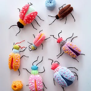 以下が含まれることがあります： 黒いワイヤーの脚と触覚が付いた、カラフルな手作りのかぎ針編みの昆虫のコレクション。昆虫は、ピンク、オレンジ、青、茶色など、さまざまな色の組み合わせが特徴です。青とオレンジの2つの毛糸玉も存在します。