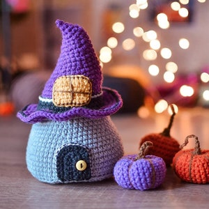 May include: A crocheted witch's house with a purple hat and a door, surrounded by three crocheted pumpkins in purple and orange.