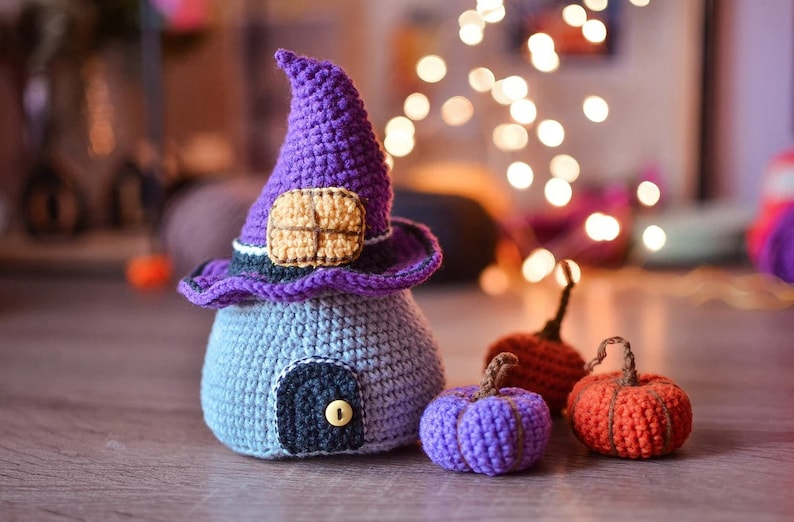 May include: A crocheted witch's house with a purple hat and a door, surrounded by three crocheted pumpkins in purple and orange.