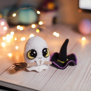 Pu&ograve; includere: Un fantasma bianco all'uncinetto con occhi grandi e un sorriso, che tiene una piccola scopa marrone. Il fantasma indossa un cappello da strega all'uncinetto nero e viola.