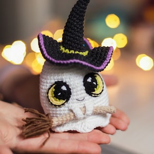 Pu&ograve; includere: Un fantasma bianco all'uncinetto che indossa un cappello da strega nero con un bordo viola e tiene una scopa marrone. Il fantasma ha occhi gialli e un piccolo sorriso.