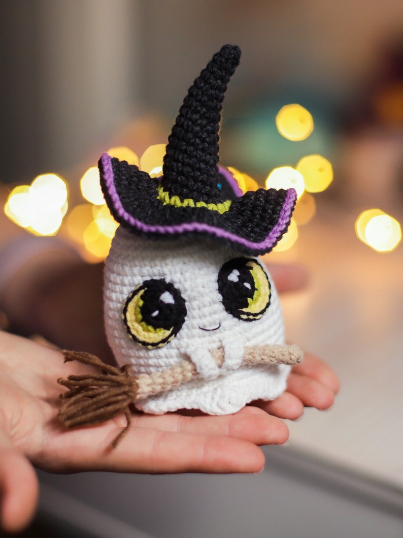 Pu&ograve; includere: Un fantasma bianco all'uncinetto che indossa un cappello da strega nero con un bordo viola e tiene una scopa marrone. Il fantasma ha occhi gialli e un piccolo sorriso.
