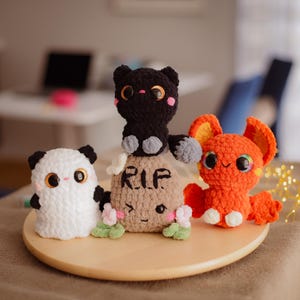 Könnte beinhalten: Ein Set aus vier gehäkelten Plüschtieren zum Thema Halloween. Eine schwarze Katze sitzt auf einem braunen Grabstein mit der Aufschrift "RIP". Ein weißer Geist und eine orangefarbene Fledermaus befinden sich auf beiden Seiten des Grabsteins.