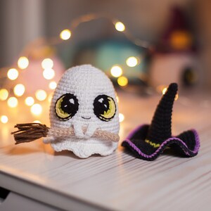 May include: A white crocheted ghost holding a brown broom with a yellow and green face and big eyes. A black crocheted witch hat with a purple brim sits next to the ghost.