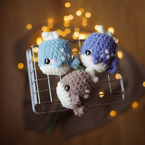 May include: Three crocheted whale plushies in a wire basket. The whales are blue, brown, and light blue with white accents. The whales are sitting in a wire basket with a blurred background of lights.