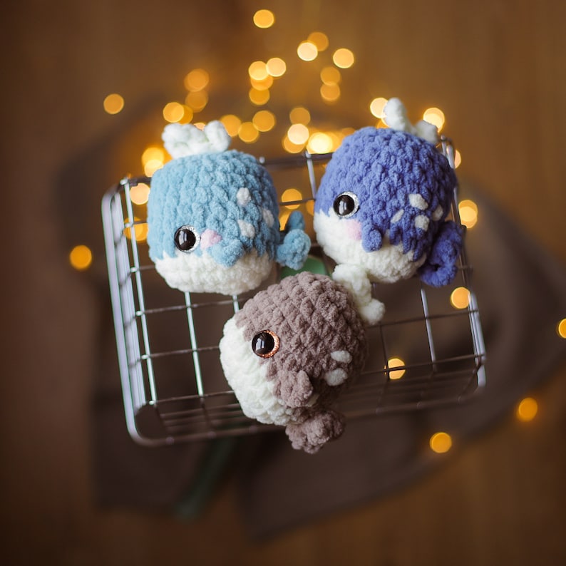 May include: Three crocheted whale plushies in a wire basket. The whales are blue, brown, and light blue with white accents. The whales are sitting in a wire basket with a blurred background of lights.
