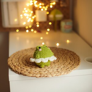 May include: A green crocheted frog wearing a white crocheted tutu sits on a woven straw mat. The frog has black eyes and a pink nose.