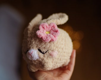 May include: A crocheted plush bunny toy with a pink flower on its head. The bunny is beige and has a white nose and black eyes.