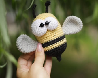 May include: A crocheted bee toy with black and yellow stripes, white eyes, and gray wings. The bee is being held by a hand.
