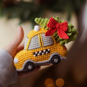 Puede incluir: Un coche de juguete de crochet amarillo con un patrón de tablero de ajedrez blanco y negro en el lateral. El coche tiene un árbol de Navidad de crochet verde y rojo en la parte superior.