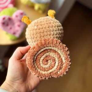 May include: A handmade, plush caterpillar toy with a light brown body and yellow antennae. The caterpillar is holding a plush, spiral-shaped roll with brown and cream colors. Other plush toys are visible in the background.