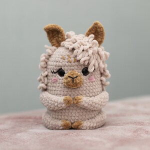 May include: A handmade, light beige crochet llama with brown accents. The llama has a fluffy mane, small ears, and a sweet expression with rosy cheeks. Its arms are folded, and it has a detailed, textured appearance.