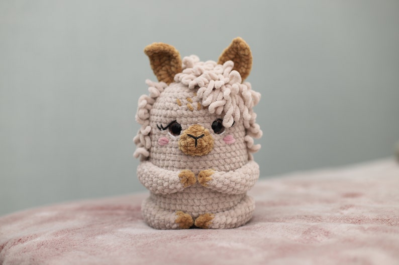 May include: A handmade, light beige crochet llama with brown accents. The llama has a fluffy mane, small ears, and a sweet expression with rosy cheeks. Its arms are folded, and it has a detailed, textured appearance.