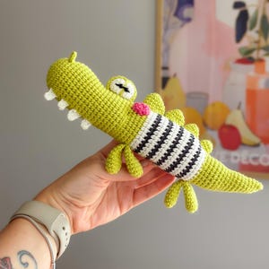 May include: A lime green crocheted alligator toy featuring a white and black striped midsection, a small pink circular detail on the neck, white triangular teeth, and two eyes with black stitched pupils, crafted with a textured yarn finish.