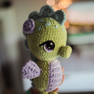 May include: A crocheted seahorse plush toy with green and purple yarn. The seahorse has a large, black eye with a purple iris and a small, purple flower on its head.
