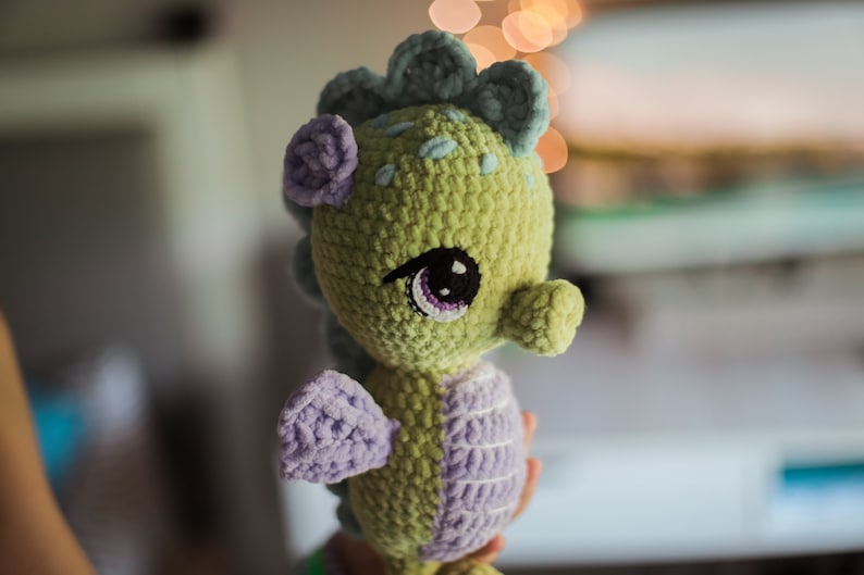 May include: A crocheted seahorse plush toy with green and purple yarn. The seahorse has a large, black eye with a purple iris and a small, purple flower on its head.