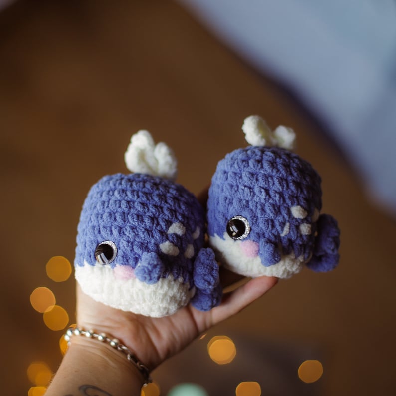 May include: Two crocheted whale plushies with white accents and black eyes. The whales are blue and have a textured surface.