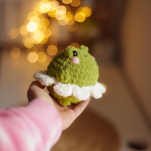 May include: A green crocheted frog wearing a white crocheted skirt. The frog has a pink nose and black eyes.