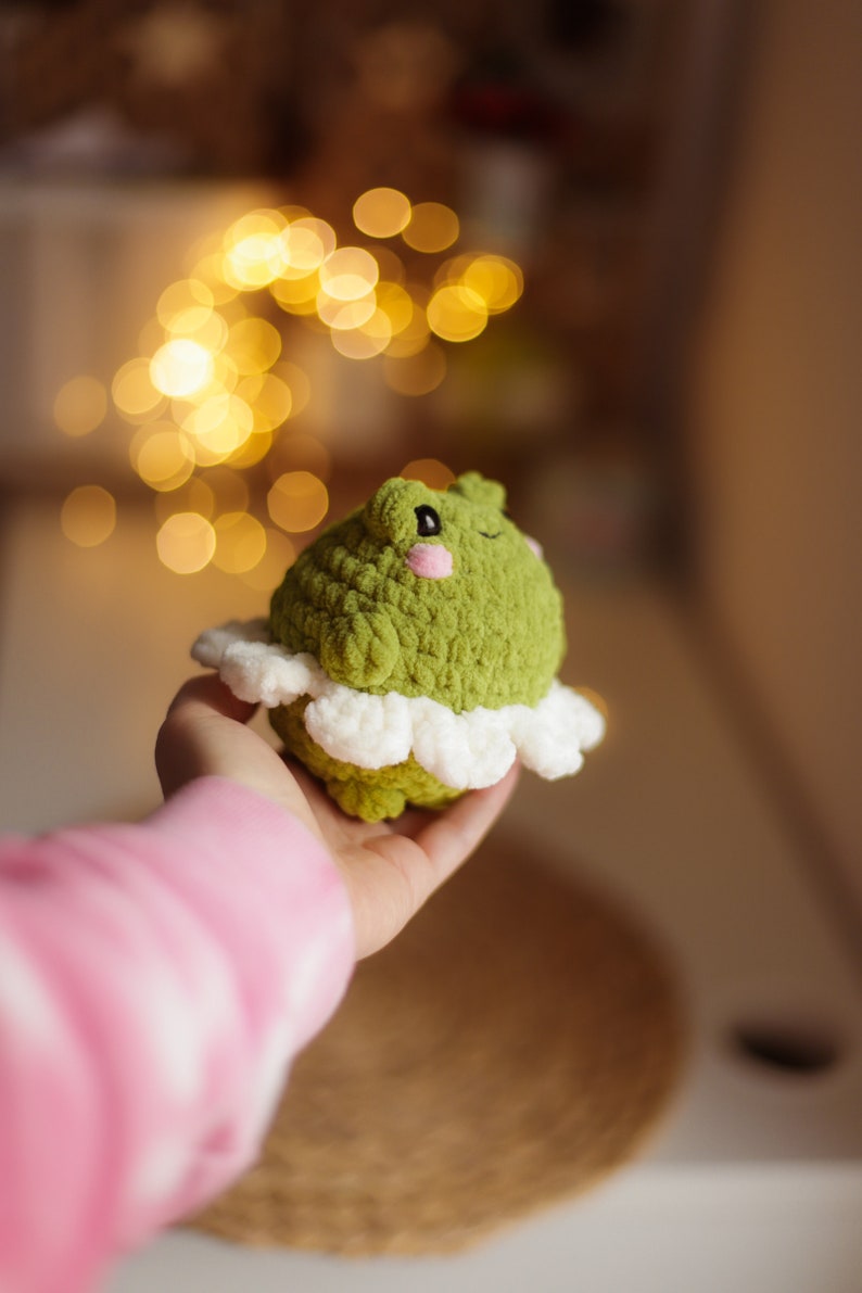 May include: A green crocheted frog wearing a white crocheted skirt. The frog has a pink nose and black eyes.