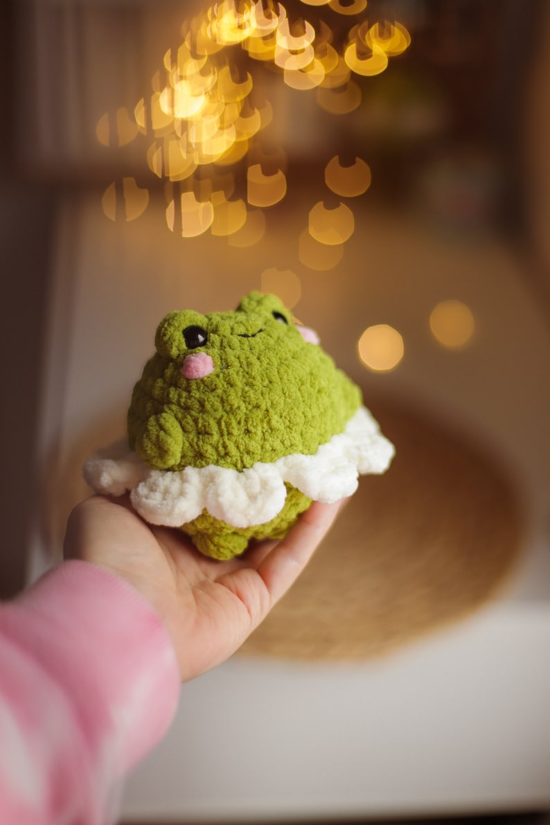 May include: A green crocheted frog wearing a white crocheted skirt. The frog has pink cheeks and black eyes. The frog is being held in a hand.