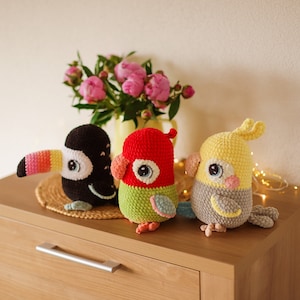 May include: Three crocheted stuffed animals: a toucan with a yellow beak and pink stripes, a red and green parrot, and a yellow and gray cockatiel. The animals are sitting on a wooden surface.