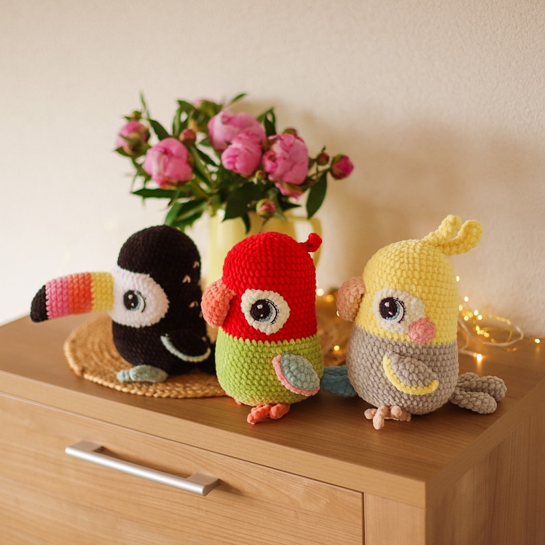 May include: Three crocheted stuffed animals: a toucan with a yellow beak and pink stripes, a red and green parrot, and a yellow and gray cockatiel. The animals are sitting on a wooden surface.