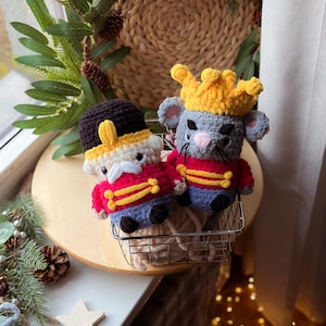 May include: Two crocheted figures: a soldier and a mouse king. The soldier wears a red jacket, blue pants, and a black hat. The mouse king has a gray body, a yellow crown, and a red jacket. They are in a small metal basket.
