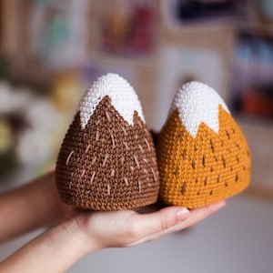 May include: Two crocheted mountain-shaped toys. One is brown with white snow peaks and the other is yellow with white snow peaks.