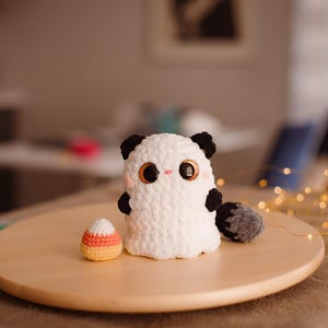 May include: A white crocheted ghost with black ears and a tail, holding a candy corn. The ghost has large, black eyes and a pink nose. The ghost and candy corn are sitting on a wooden surface.